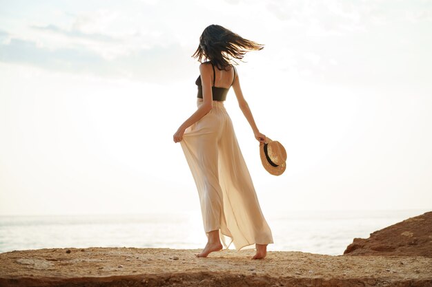 Young stylish woman standing on cliff at the sea at sunset