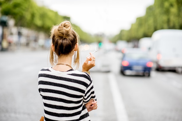 Foto giovane donna elegante in maglione a righe con occhiali fumando una sigaretta in piedi all'aperto per strada a parigi