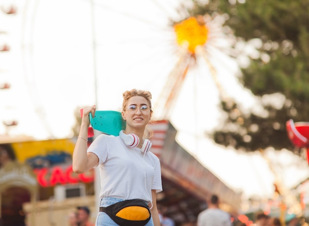 Young stylish woman in stylish youth clothes with a skateboard\
on her shoulders having fun in amusement park