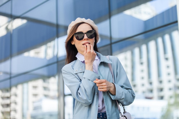 Young stylish woman in sunglasses and beret paints her lips against the background of the windows of the business center Fashion beauty lifestyle