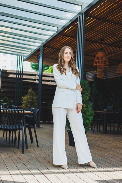 A young stylish woman in a white classic suit with a smile on her face is standing on the terrace of a summer cafe