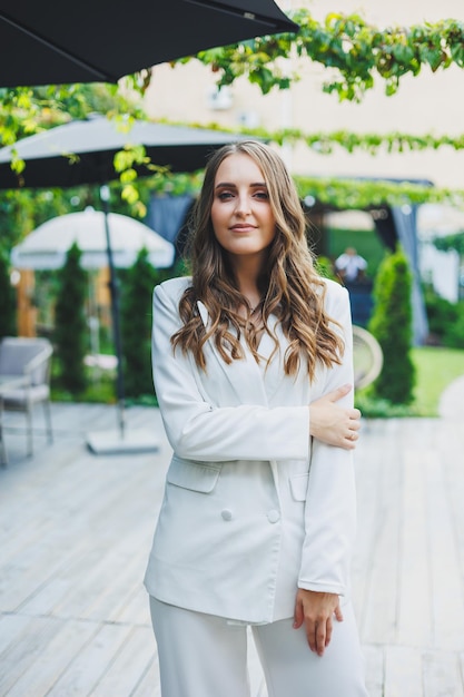 A young stylish woman in a white classic suit with a smile on her face is standing on the terrace of a summer cafe