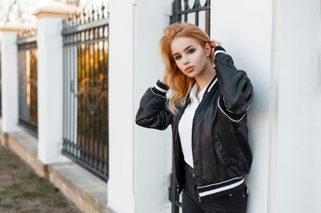 Young stylish woman with beautiful eyes in a black summer stylish jacket in black jeans in a white polo shirt posing against a vintage metal fence. Nice modern girl.