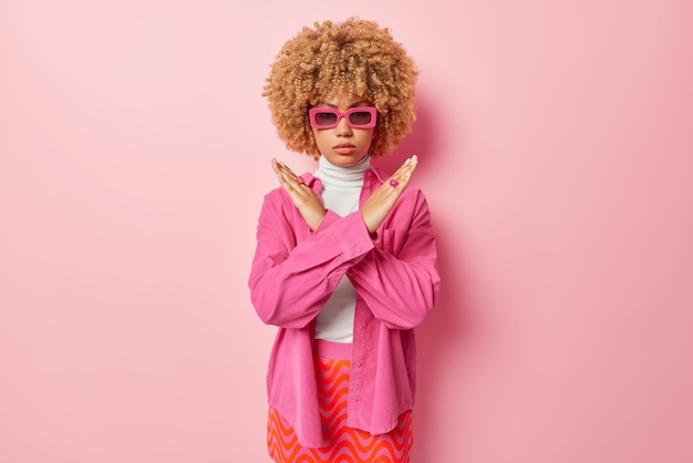 Young stylish woman with curly hair makes taboo gesture crosses\
arms denies something asks to stop wears trendy sunglasses shirt\
and skirt stands against pink background body language concept