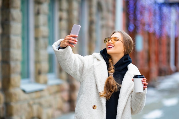 Giovane donna alla moda con smartphone e caffè all'aperto fa un selfie