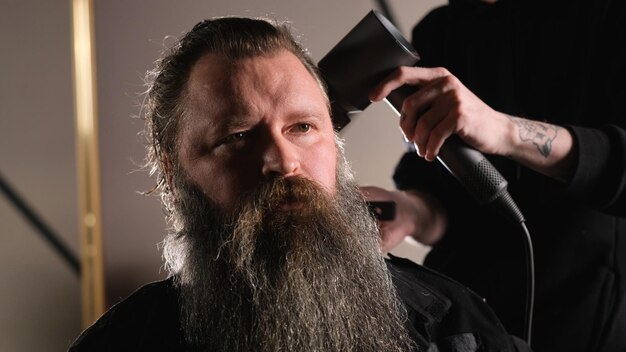 A young stylist does styling with a hair dryer to a bearded man sitting in a barber chair
