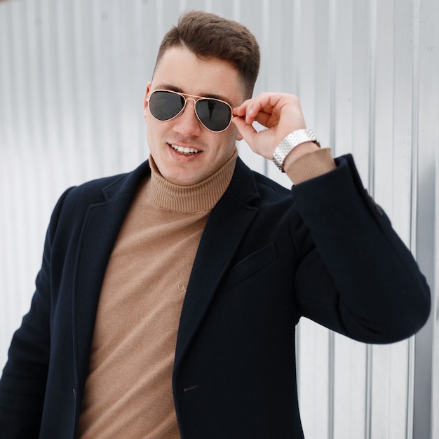 Young successful attractive male businessman in stylish sunglasses in an elegant coat in a knitted sweater with a silver clock