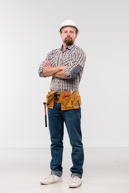 Young successful bearded repairman in white hardhat, checkered shirt and blue jeans standing in front of camera in isolation