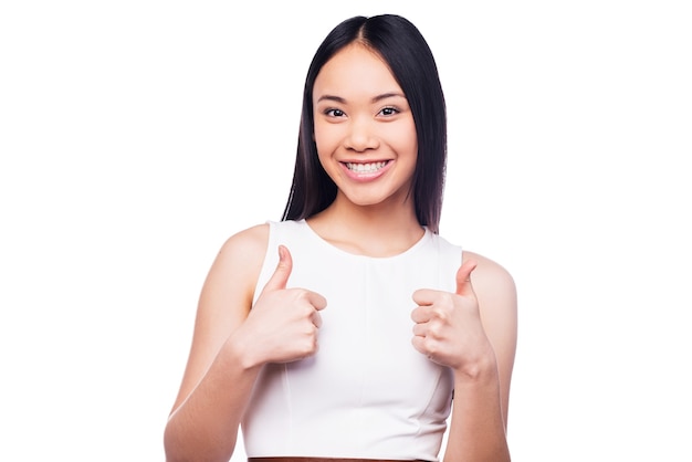 Молодой и успешный. Красивая молодая азиатская женщина смотрит в камеру и показывает палец вверх, стоя на белом фоне