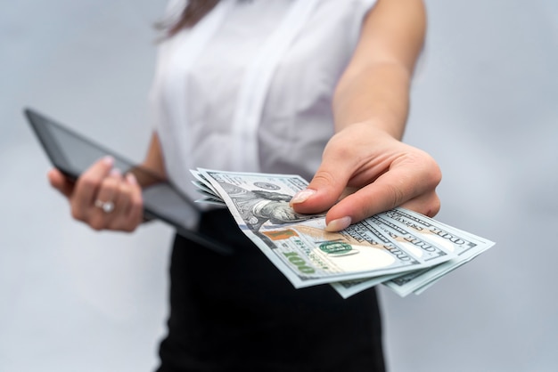 Young successful business woman holding dollar bills and using tablet isolated grey background.