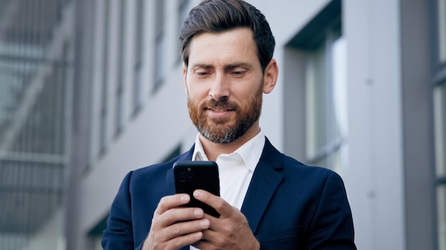 Young successful businessman holding smartphone standing in city business center uses phone writes