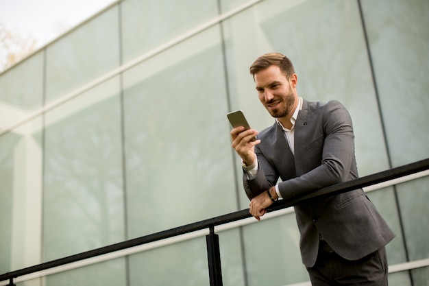 Young successful businessman wearing grey suit and holding his smartphone while standing near modern