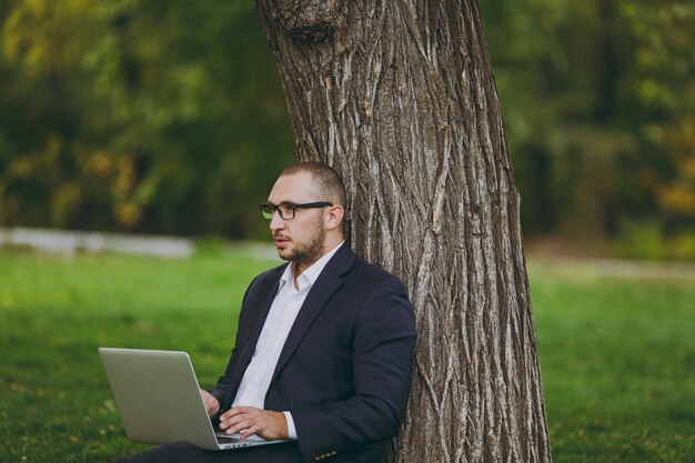 Young successful businessman in white shirt, classic suit, glasses. Man sit on grass ground, work on laptop pc computer in city park on green lawn outdoors on nature. Mobile Office, business concept.