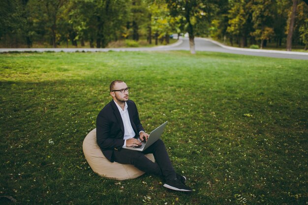 Young successful businessman in white shirt, classic suit, glasses. Man sit on soft pouf, working on laptop pc computer in city park on green lawn outdoors on nature. Mobile Office, business concept.