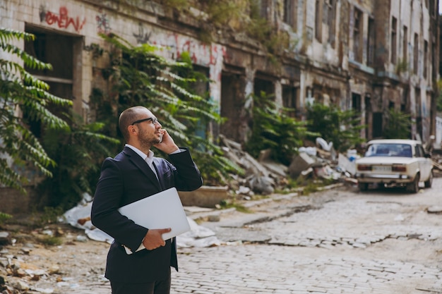 Young successful businessman in white shirt, classic suit, glasses. Man standing with laptop pc computer, talking on phone near ruins, debris, stone building outdoors. Mobile Office, business concept.