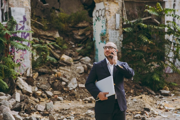 Young successful businessman in white shirt, classic suit, glasses. Man standing with laptop pc computer, talking on phone near ruins, debris, stone building outdoors. Mobile Office, business concept.