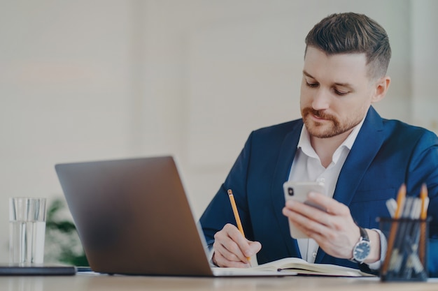 Young successful businessman with mobile phone in hand writing down important data into notepad while sitting in front of laptop at office, wearing stylish dark blue formal suit and expensive watch