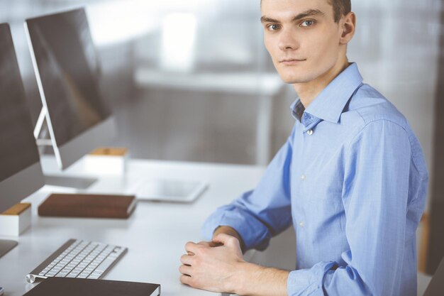 Photo young successful businessman works with computer, sitting at the desk in a modern office. headshot of male entrepreneur or it-specialist at workplace. business concept