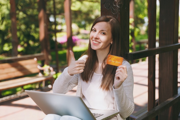 Young successful businesswoman holding credit card. Woman sitting on bench working on modern laptop pc computer in city park in street outdoors on nature. Mobile Office. Freelance, business concept.