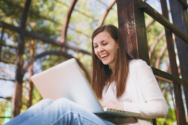 Young successful businesswoman in light casual clothes. Smiling Woman working on modern laptop pc computer in city park in street outdoors on spring nature. Mobile Office. Freelance business concept.