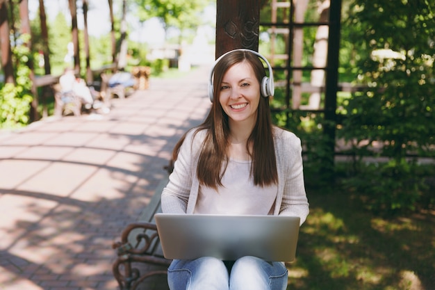 Young successful businesswoman in light casual clothes. Woman working on modern laptop pc computer, listen music in headphones on head in street outdoors. Mobile Office. Freelance business concept.