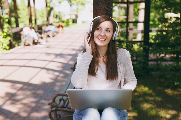 Young successful businesswoman in light casual clothes. Woman working on modern laptop pc computer, listen music in headphones on head in street outdoors. Mobile Office. Freelance business concept.