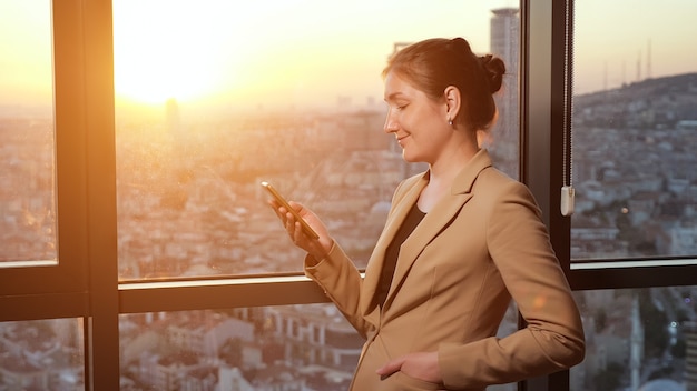 Young successful businesswoman in office suit is browsing mobile phone, side view. She is standing near the window with panoramic city view in her office in evening, sunlight