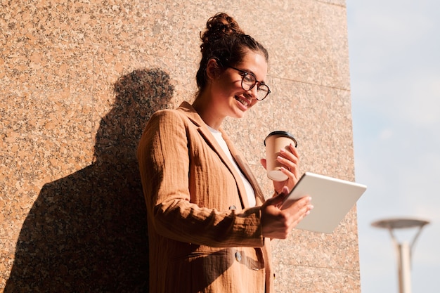 Young successful businesswoman in smart casualwear having glass of coffee and using digital tablet against wall of building outdoors