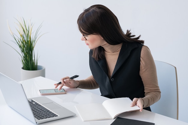 Young successful businesswoman with open copybook scrolling in smartphone while sitting by desk in front of laptop