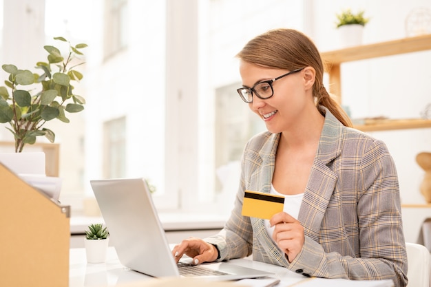 Young successful businesswoman with plastic card looking at laptop display while searching through online goods