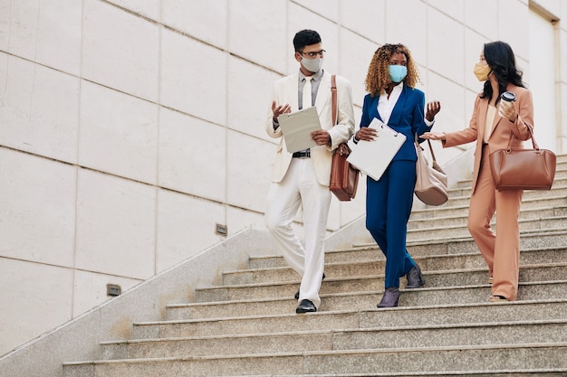 Young successful busness people in protective masks walking down stairs to office and talking about development of recent project