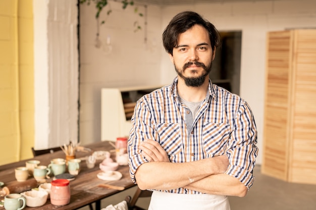 Young successful cross-armed master of pottery standing on workplace