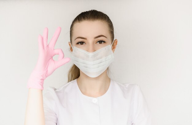 Young successful doctor in protective mask and white coat shows sign with okay hand