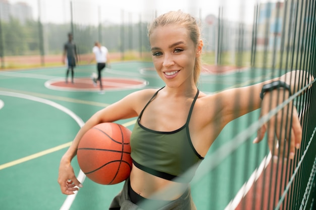 Giovane giocatore di pallacanestro femminile di successo con la palla che ti guarda mentre fa una pausa rete sul parco giochi all'aperto