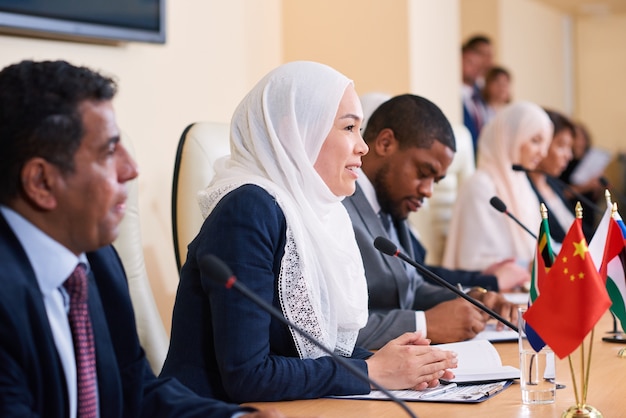Молодая успешная женщина-делегат в хиджабе принимает участие в обсуждении отчета коллеги во время разговора в микрофон на конференции