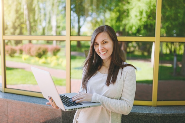 Young successful girl in light casual clothes. Beauty woman working on modern laptop pc computer near mirror building with tree reflection in street outdoors. Mobile Office. Freelance business concept