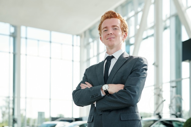 Молодой успешный мужчина-менеджер автоцентра смотрит на вас, стоя против новых автомобилей перед камерой и ожидая клиентов