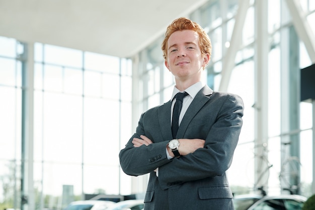 Young successful male manager of auto center looking at you while standing against new cars in front of camera and waiting for customers