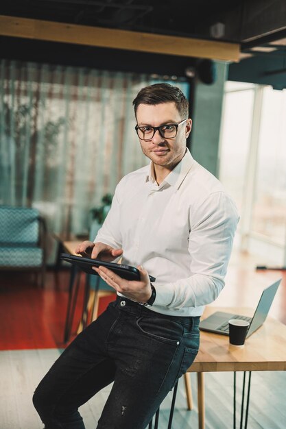 Foto un giovane di successo con gli occhiali e una camicia bianca sta lavorando su un tablet su un progetto in uno spazio di lavoro moderno un uomo d'affari, direttore, lavora in ufficio