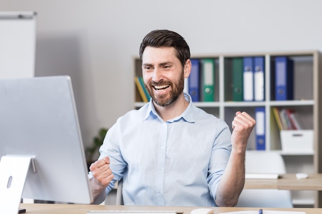 Young successful man office worker manager freelancer rejoices\
in the result of the work done he sits happy at the table and\
computer rejoices raises his hands