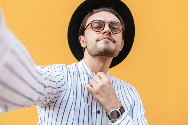 Фото Молодой успешный человек в очках и черной шляпе делает селфи-фото и показывает наручные часы, изолированные на желтой стене