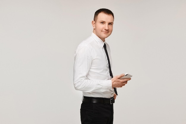 A young successful man in a white shirt and a black tie keeps the phone in his hand on a gray backgr...