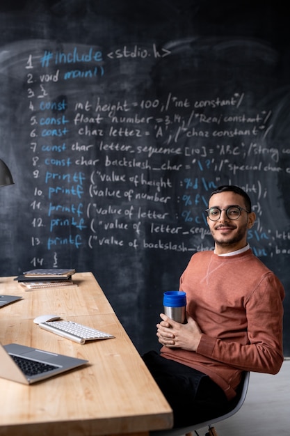 Giovane programmatore di successo con la tazza che ti guarda mentre era seduto alla scrivania sopra la lavagna con formula scritta