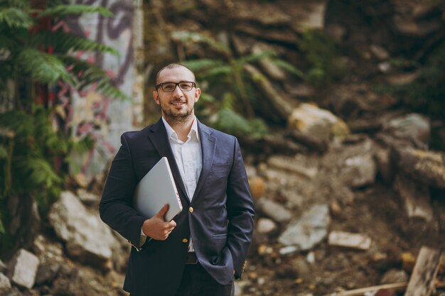 Young successful smart businessman in white shirt, classic suit, glasses. Man standing with laptop pc computer phone near ruins, debris, stone building outdoors. Mobile Office, business, work concept.