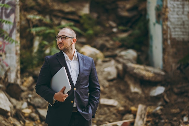 Photo young successful smart businessman in white shirt, classic suit, glasses. man standing with laptop pc computer phone near ruins, debris, stone building outdoors. mobile office, business, work concept.