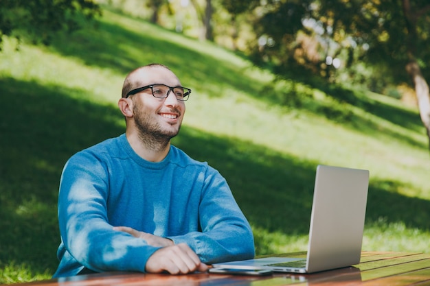 Giovane uomo d'affari o studente sorridente intelligente di successo dell'uomo felice in camicia blu casual, occhiali seduti al tavolo con il telefono cellulare nel parco cittadino utilizzando il computer portatile, lavorando all'aperto. concetto di ufficio mobile.