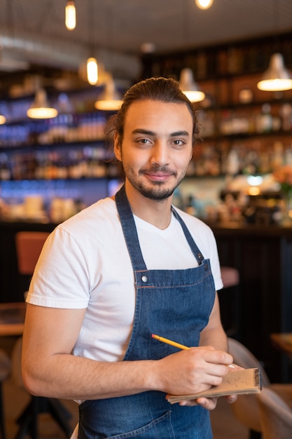 Young successful waiter in apron making notes in notepad while taking order of client during work in cafe or restaurant