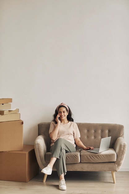 Young successful woman sitting on sofa with laptop she accepting orders by the phone with big stacks of parcels near the sofa prepared for delivering