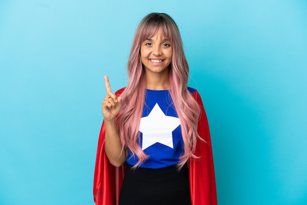 Молодая супергеройская женщина с розовыми волосами изолирована на синем фоне, указывая на отличную идею