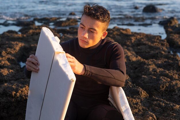 Young surfer sitting on the rocks looks thoughtfully at the broken board in half Handsome millennial guy with wetsuit Horizon over the water and sunset light